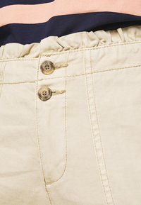 Beige kraťasy s nařaseným elastickým páskem, které mají dva hnědé knoflíky a prošívané detaily. Hladká, odolná tkanina s jemnou strukturou.