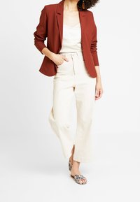Blazer couleur rouille sur un haut clair, associé à un pantalon large crème et des sandales à motifs. Tissu doux, design sur mesure.
