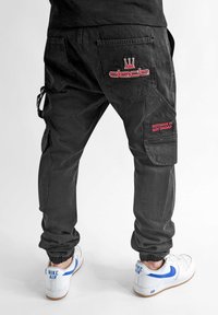 Schwarze Cargo-Hosen mit elastischen Knöchellaschen, versehen mit gestickten roten Logos auf den hinteren Taschen und einer seitlichen Cargo-Tasche.