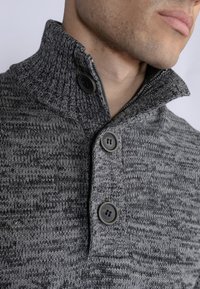 Maglione grigio a maglia con colletto alto, tre bottoni neri e un motivo testurizzato su tutta la superficie. La vista ravvicinata mette in risalto i dettagli della cucitura.