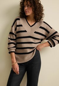 Beige und schwarz gestreifter Strickpullover mit V-Ausschnitt, oversized Passform und gerippter Textur. Kombiniert mit schwarzen Hosen, die einen lässigen Stil zeigen.