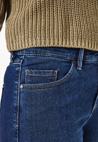 Dunkelblaue Jeans aus Denim mit einem figurbetonten Schnitt, die über eine aufgesetzte Tasche und kontrastierende orangefarbene Nähte verfügen. Ein gestrickter, beigefarbener Pullover ist teilweise sichtbar.