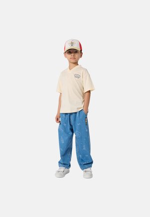 Jeune garçon portant une chemise à manches courtes beige clair, un jean ample bleu à motifs, des baskets blanches et une casquette blanche et rouge avec des motifs colorés.