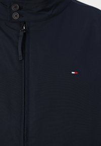 Veste bleu marine avec col montant, fermeture éclair et deux boutons noirs. Petit logo brodé en rouge, blanc et bleu.