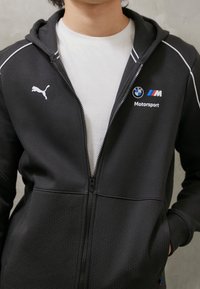 Sudadera negra con cremallera hecha de tela suave con secciones de malla, que presenta un logo blanco de Puma y la marca BMW Motorsport en el pecho.