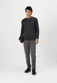 Jersey gris oscuro de cuello redondo con un pequeño logo amarillo en el pecho, combinado con jeans grises y zapatillas negras. El modelo sostiene un bolso negro.