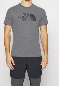 Camiseta de algodón gris con un logo negro de "The North Face" en un diseño curvado en el pecho. Cuello redondo clásico y mangas cortas.