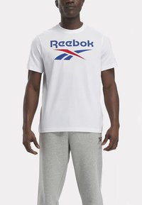 Mann i en hvit Reebok T-skjorte med en blå og rød logo, og lysegrå Reebok joggebukser, stående mot en ensfarget bakgrunn.
