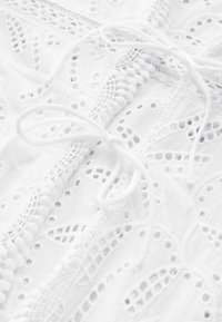 Witte stof met ingewikkelde uitgesneden borduursels, zachte textuur en decoratieve lintstrikken. Beschikt over een kantachtig patroon en pomponaccenten.