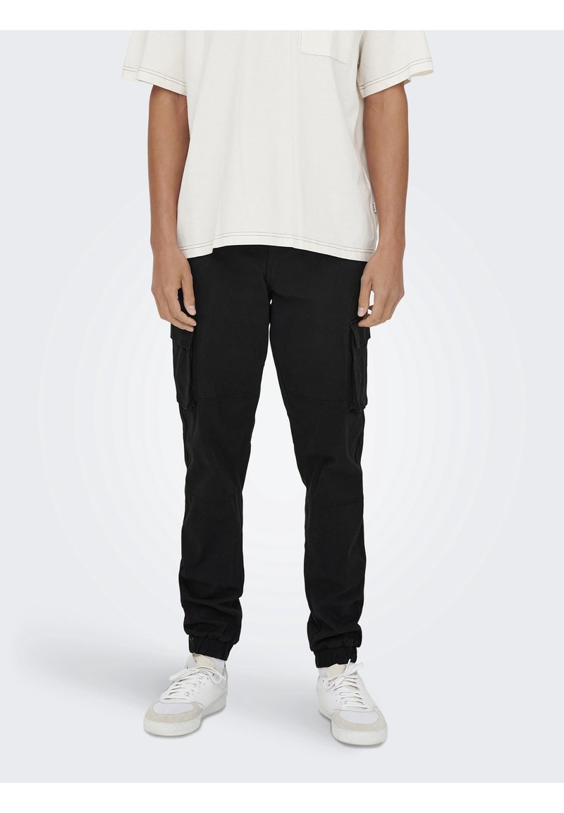 Personne portant un pantalon jogger cargo noir avec des poignets élastiques, des baskets blanches et un t-shirt blanc ample à manches courtes, debout devant un fond uni.
