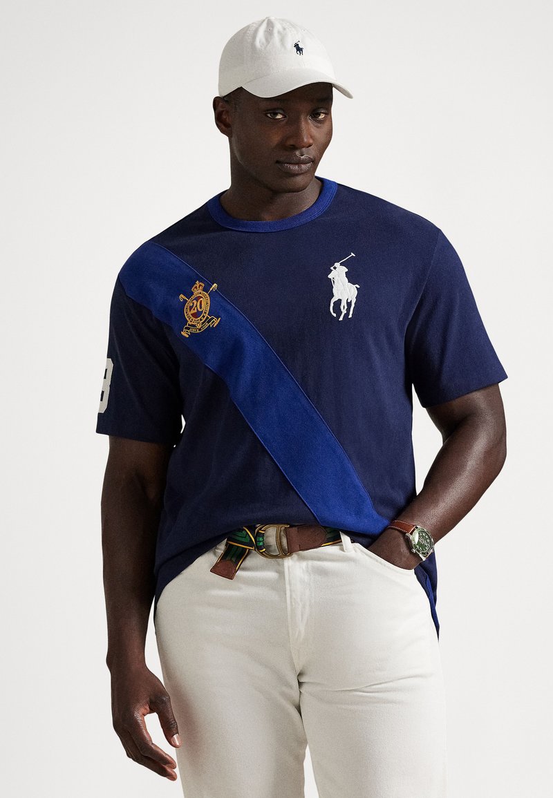 Μπλε σκούρο μπλουζάκι πόλο με κοντά μανίκια, με αντίθετη μπλε διαγώνια ρίγα, κεντημένο λογότυπο και αριθμό στο μανίκι. Συνδυασμένο με λευκό παντελόνι.