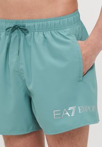 Uimaporten vaaleassa teal-värissä, valmistettu sileästä kankaasta. Sisältää joustavan vyötärönauhan, jossa on nauhakuja, sekä hopea logo-printin.