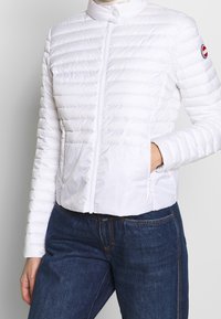 Vit puffjacka med dragkedja fram, ståkrage, horisontell sömnad och två sidofickor. Har varumärkeslogotyp på vänstra ärmen.