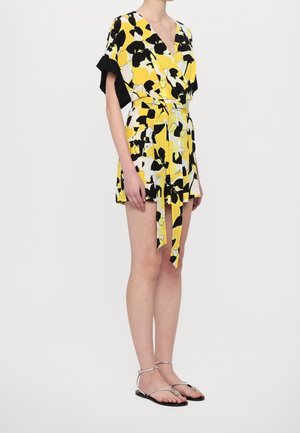 Kvinde iført en gul, sort og hvid kort kjole med blomsterprint, bælte og sølvfarvede flade sandaler på en ensfarvet baggrund.