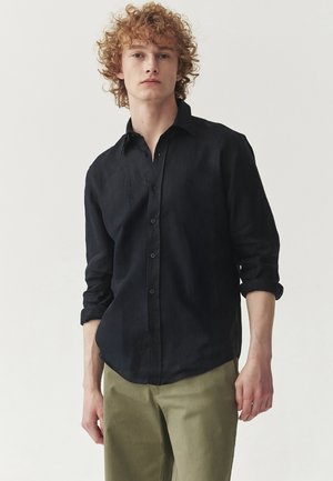 Black linen button-up shirt with long sleeves, rolled cuffs, and a relaxed fit, paired with light olive green pants. Plain design, no patterns.