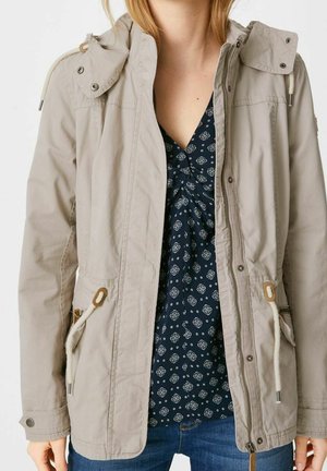 Jasnobeżowa kurtka z kapturem i sznurkami noszona na granatowej wzorzystej bluzce i niebieskich dżinsach na osobie.