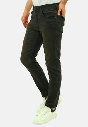 Man die strakke zwarte jeans draagt met handen in de zakken en witte sneakers, staand tegen een effen lichte achtergrond.