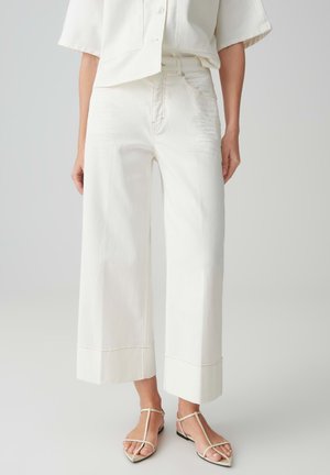 Frau, die weiße, weit geschnittene, cropped Hosen, ein weißes Kurzarmhemd und beige T-Riemchen flache Sandalen trägt, vor einem einfarbigen Hintergrund stehend.