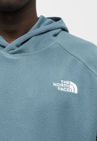 Μπλε φλις φούτερ με τσέπη καγκουρό, με λευκό λογότυπο «The North Face» στο αριστερό στήθος και κουκούλα με κορδόνια.