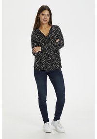 Schwarze Bluse mit weißen Punkten, langen Ärmeln und V-Ausschnitt, kombiniert mit dunkelblauen Jeans und weißen Sneakers, vor einem weißen Hintergrund.