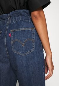 Jeans med elastisk midja, mörkblå färg och en distinkt bakfickdesign med gyllene stygn och röd etikett.