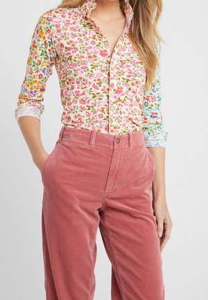 Femme portant une chemise à fleurs multicolores boutonnée avec les manches retroussées et un pantalon en velours côtelé rose taille haute, debout avec une main dans la poche.