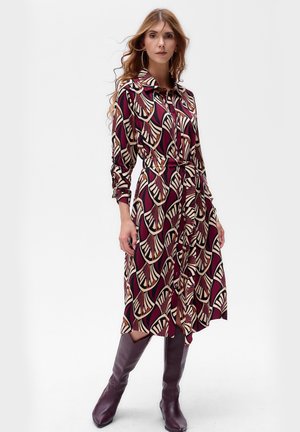 Kleid mit Kragen in Maroon mit einem Fächer-Muster in Creme und Schwarz. Lange Ärmel, tailliert und knielanger Saum. Kombiniert mit knielangen lila Stiefeln.