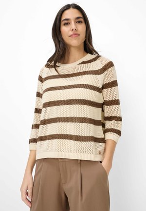 Frau trägt einen beigen Strickpullover mit braunen horizontalen Streifen und Dreiviertelärmeln, kombiniert mit braunen hoch taillierten Hosen.