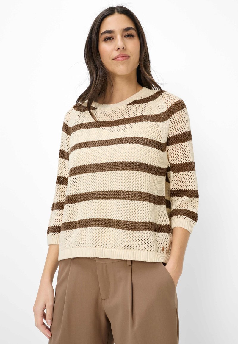 Frau trägt einen beigen Strickpullover mit braunen horizontalen Streifen und Dreiviertelärmeln, kombiniert mit braunen hoch taillierten Hosen.
