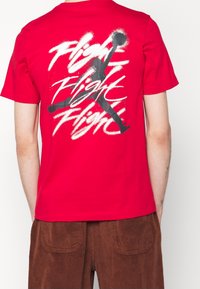 Punainen puuvillapaita, jossa on suuri, kontrastivärinen musta graafinen suunnittelu, jossa sana "Lento" on toistettu useita kertoja ja siluetti-ikoni.