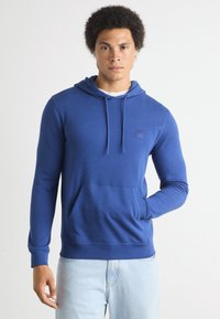 Felpa blu realizzata in stoffa morbida, con tasca a marsupio, cordini regolabili e un sottile patch con logo sul petto.