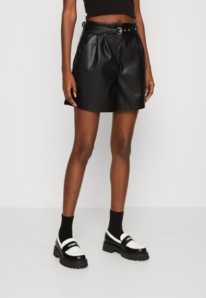 Person iført sort lædershorts med høj talje og bælte, sorte sokker og chunky sorte-hvide loafers, stående mod en ensfarvet hvid baggrund.