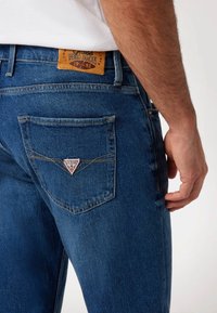 Liemenės mėlynos džinsinės kelnės turi odinę prekės etiketę ir trikampio logotipą ant galinio kišenės, su kontrastingais siuvinėjimais.