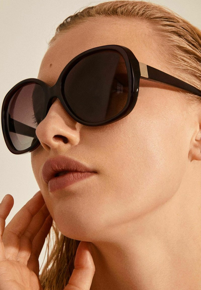Occhiali da sole neri oversize con lenti sfumate, ampia montatura in plastica e dettaglio metallico sulle tempie, indossati da una persona con pelle chiara.