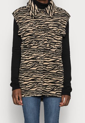 Personne portant un gilet sans manches à motifs zébrés boutonné sur un haut noir à manches longues et un jean bleu, debout devant un fond blanc.