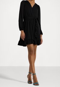 Svart omlottklänning med långa ärmar, volangkant och midjeband. Bärs med turkosa ankelsandaler mot en enkel vit bakgrund.