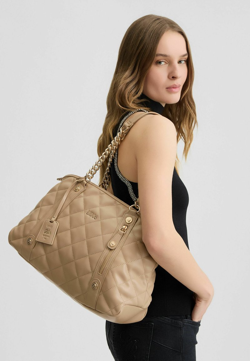 Borsa a tracolla beige trapuntata con catena e dettagli in metallo dorato, dotata di chiusura con zip e logo sul davanti.