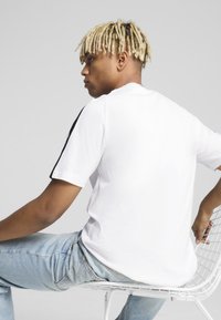Camiseta blanca de manga corta con franjas laterales negras, hecha de algodón. Modelo sentado en una silla de alambre, vistiendo jeans de color azul claro.