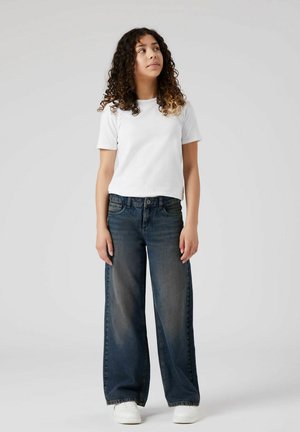 Camiseta blanca de manga corta combinada con vaqueros de pierna ancha azul oscuro. Los vaqueros tienen un lavado descolorido, bolsillos delanteros y cierre de botón. Zapatillas blancas.