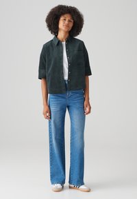 Dark green corduroy short-sleeve shirt with a button front and chest pocket, paired with light blue wide-leg jeans and white sneakers.