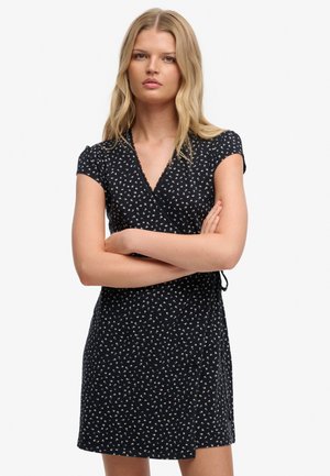 Jonge vrouw met lang blond haar draagt een zwarte wikkeljurk met korte mouwen en een klein wit bloemenpatroon, armen gekruist.