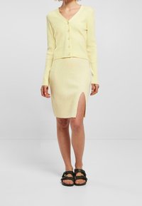 Cardigan giallo a costine con scollo a V e bottoni, abbinato a una gonna abbinata di lunghezza al ginocchio con uno spacco laterale. Sandali neri esposti.