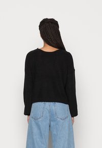 Pull en maille côtelée noire avec un large décolleté et des manches longues, associé à un jean taille haute bleu clair, mettant en valeur la texture et la coupe.