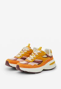 Zapatillas de ante en naranja y marrón con detalles de malla, cordones blancos y una suela gruesa. Presentan una lengüeta de tirón y un logotipo sutil en la lengüeta.