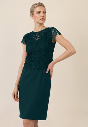 Frau trägt ein dunkelgrünes Spitzen-Top mit kurzen Ärmeln und einen dazu passenden, eng anliegenden Bleistiftrock, posiert mit einer Hand in der Hüfte und schaut seitlich.