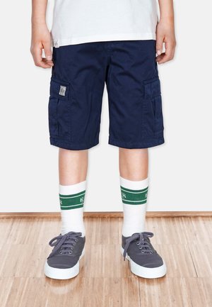 Marine Cargo-Shorts mit Seitentaschen, kombiniert mit weißen Socken mit grünen Streifen und grauen Sneakers mit weißen Sohlen.