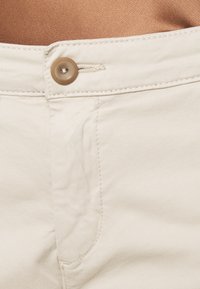 Pantalones de algodón beige con cierre de botón en la parte delantera, con costuras visibles y una textura suave. La tela tiene un brillo sutil.