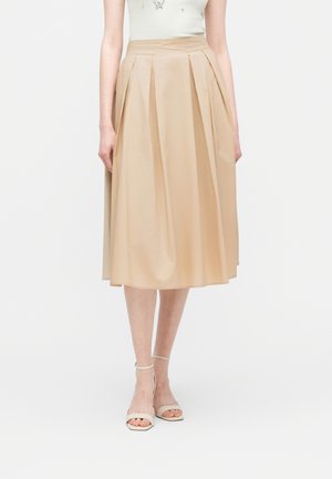 Naine, kes kannab beežikujulist midi-seelikut esi voltidega, kombineeritud valgete kõrge kontsaga pahkluu rihmaga sandaalidega, ühetaustal.