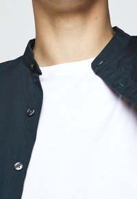 Ciemnozielona koszula z mandaryńskim kołnierzykiem, z czarnymi guzikami. Nosi się ją na gładką białą koszulkę.