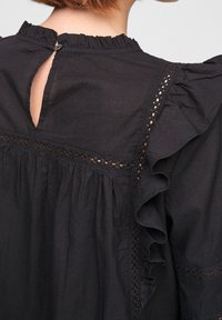 Schwarze Bluse mit Rüschenausschnitt, Schlüsselrücken mit Knopfverschluss und Spitzenakzenten entlang der Nähte und Ärmel.
